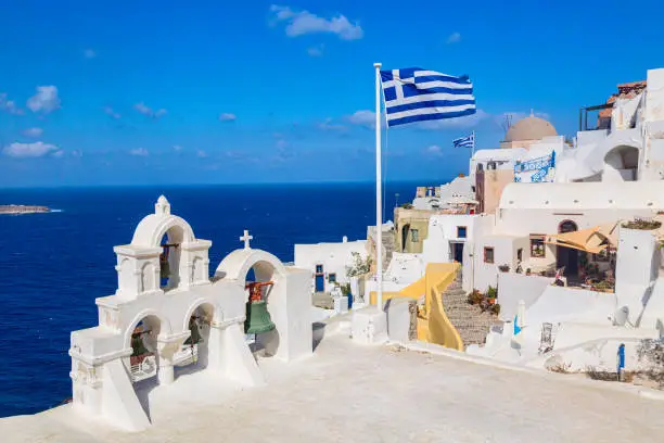 Photo of Greek orthodox church with bells and greek flag against famous white houses on Santorini island, Aegean sea, Greece.