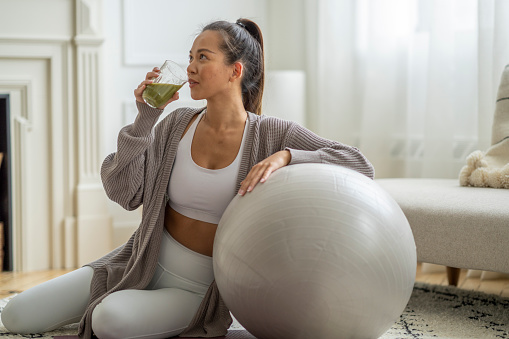 A young woman of Asian decent, poses for a portrait before starting a fitness regimen at home.  She is wearing comfortable athletic wear and is sitting with her arm resting on a large yoga ball and a green smoothie in the other hand. 
 She is drinking the smoothie before beginning her workout.