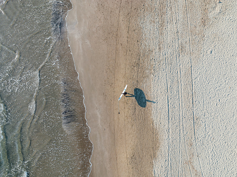 Aerial view of a man walking at the beach with his surfboard. His shadow is visible on the sand.