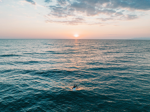 Aerial view of a man surfing in the sea at sunset. He's swimming with his surfboard.