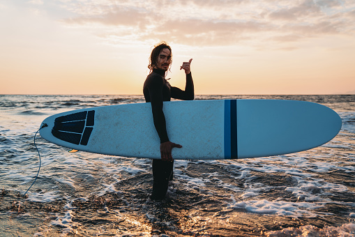 Portrait of a man with his surfboard standing in the sea making the shaka sign. He's looking at camera. Sunset time.