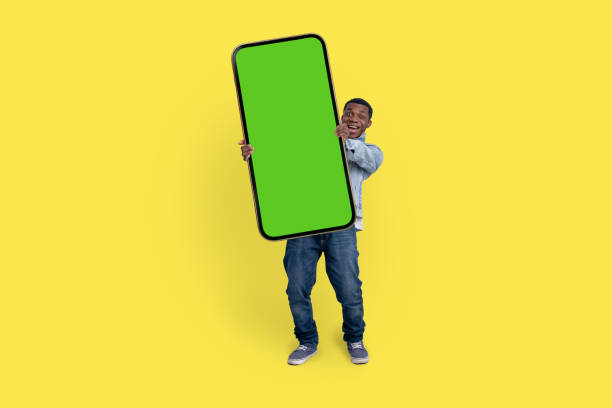 Excited black man standing and pointing Big smartphone with blank green screen, demonstrating copy space for app or website design, standing over yellow studio background, mockup image stock photo