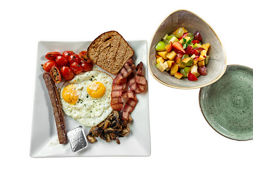 Breakfast plate isolated on white background. Clipping path included. Healthy eating at morning with egg, sausage, bacon, mushroom, tomato, bread and fruit salad.