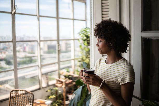 giovane donna contemplativa che tiene una tazza e guarda attraverso la finestra di casa - guardare fuori foto e immagini stock