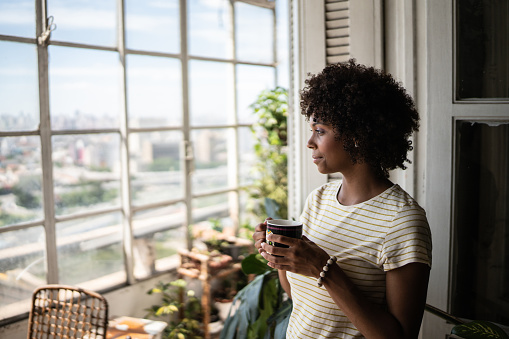 Contemplative young woman holding a mug and looking through the window at home