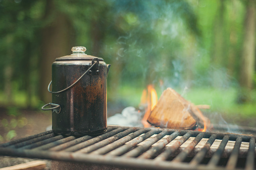 A pot of water warms up on a metal grate over a fire at a campground in the summer.