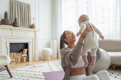 A new mother of Asian decent, sits on the floor of her living room with her baby in her arms as she works out at home with the infant.  She is dressed comfortably in athletic wear and is seated on a yoga mat as she exercises with her baby.