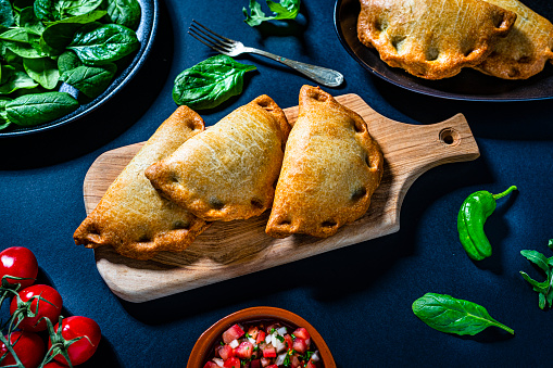 High angle view of a group of fresh spicy Argentinian empanadas on a cutting board shot on dark table. High resolution 42Mp studio digital capture taken with Sony A7rII and Sony FE 90mm f2.8 macro G OSS lens