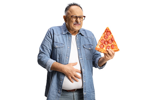 Mature man holding a slice of pepperoni pizza and holding his abdomen in pain isolated on white background