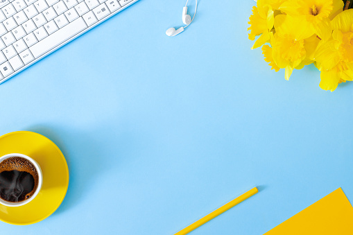 Woman's modern workspace. Keyboard, glasses and office supplies on blue table. Bouquet of delicate beautiful yellow daffodils and cup of coffee for holiday. 
Flat lay  top view  concept with copy space for text.
