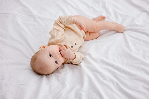 A cute blue-eyed six-months-old baby laying in thoughts on white linen on the bed looking at camera sucking thumbs