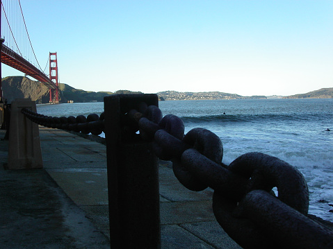 View of Golden Gate bridge from Fort Point during sundown. Surfers hidden in the waves. Pavement wet from ocean spray.