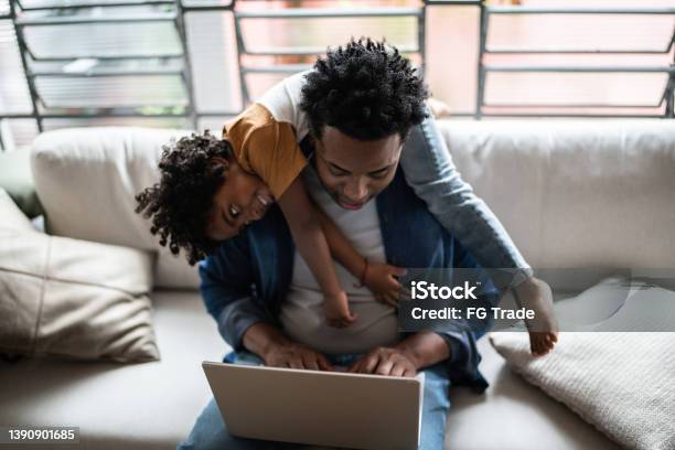 Father Using The Laptop Trying To Work While Son Is On His Back At Home Stock Photo - Download Image Now