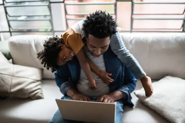 Photo of Father using the laptop trying to work while son is on his back at home