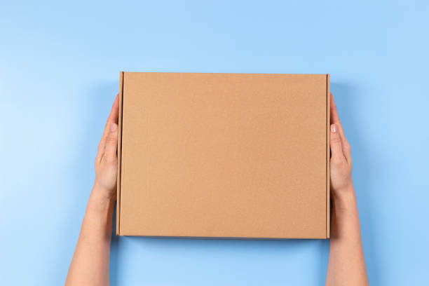 istock Top view to female hands holding brown cardboard box on light blue background. Mockup parcel box. Packaging, shopping, delivery concept 1390904410