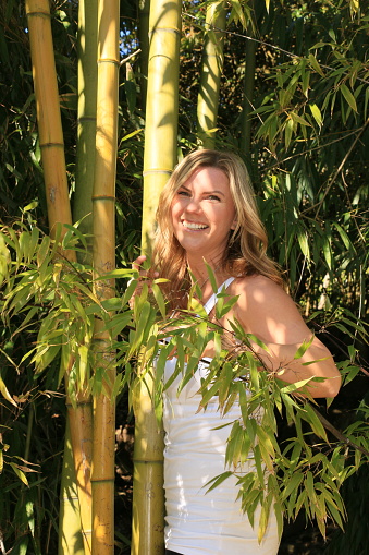 A Caucasian woman in a stand of Bamboo. She is wearing a white sleeveless top.
