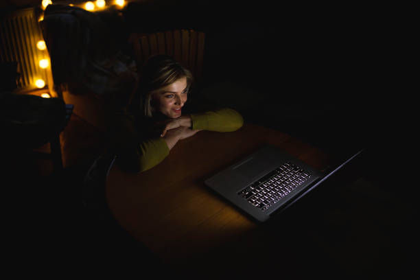 High angle view of happy mid adult woman sitting on the floor in her living room, resting head and hands on the coffee table and enjoying watching a fun movie on laptop, late at night.