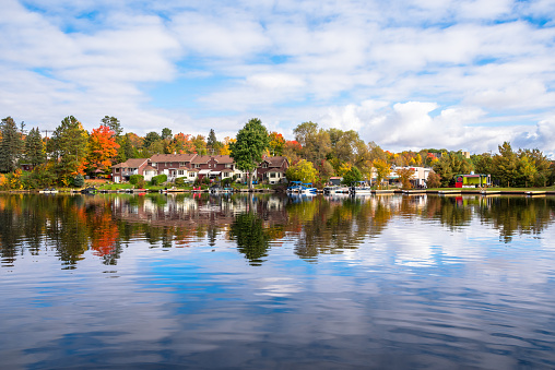 View of modern lakeside brick row houses surrounded by colourful trees at the peak of fall foliage on a partly cloudy morning. Reflection in water. Huntsville, ON, Canada.