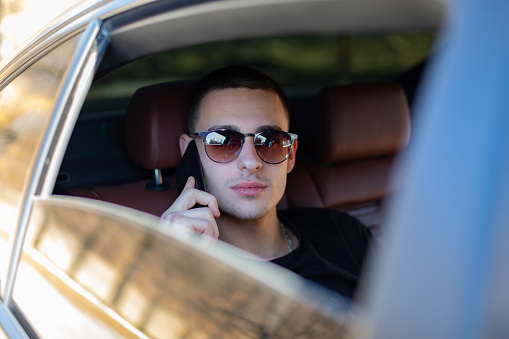 Young man sitting on a back seat of a car using his self phone on the road, making plans, searching, surfing the internet and media, talking , smiling and enjoing road trip ona a beautiful sunny day.