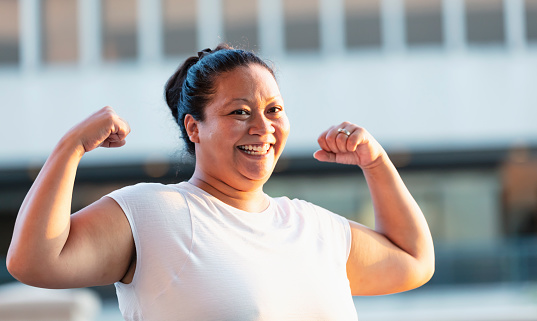 Headshot of a Pacific Islander woman in her 30s standing outdoors in the city, flexing her biceps, grinning at the camera.