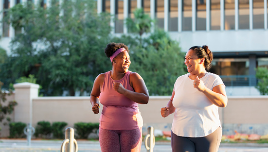 Two multiracial women exercising in the city. They are mature, plus size women, staying in shape together, running, jogging or power walking side by side. The African-American woman is in her 40s. Her Pacific Islander friend is in her late 30s.