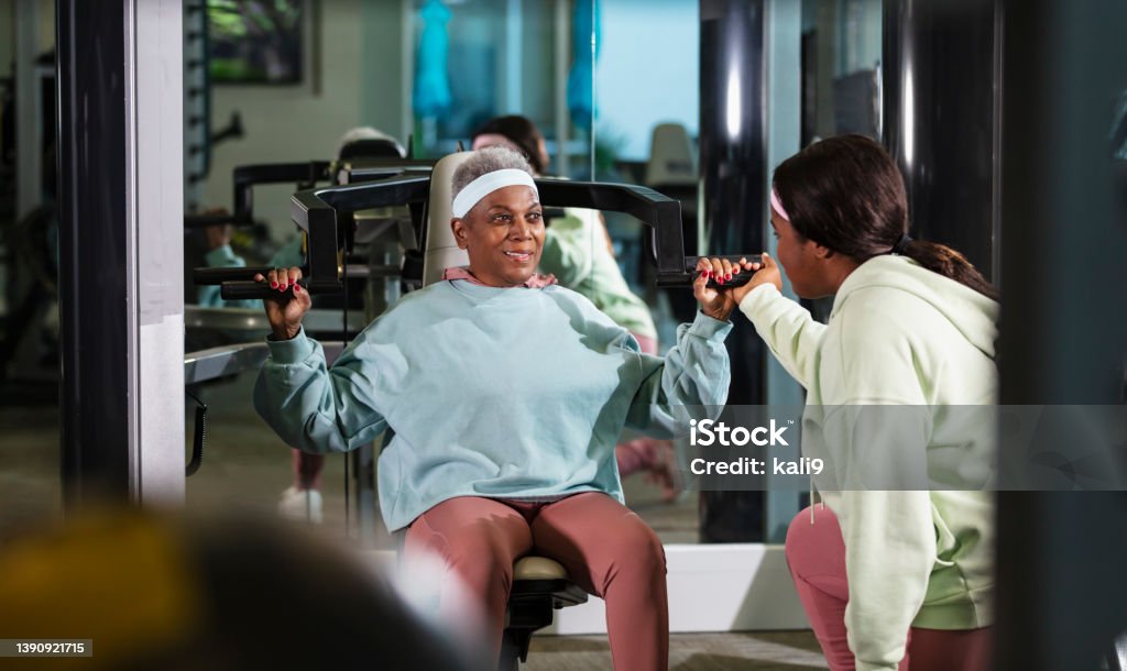 Senior black woman at gym smiling at personal trainer A senior African-American woman, in her 70s, at the gym exercising, using a shoulder press machine to strengthen her deltoids. A young woman is her fitness instructor, assisting her with her exercises. 25-29 Years Stock Photo