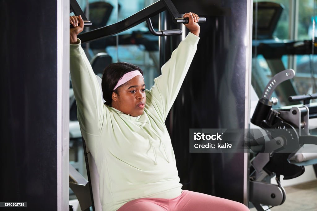 African-American woman using shoulder press at gym A young African-American woman, in her 20s, at the gym exercising, using a shoulder press machine to strengthen her deltoids. . 25-29 Years Stock Photo