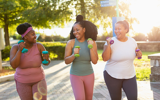 A multiracial group of three women exercising together in a city park. They are power walking with hand weights, side by side, smiling and conversing. The two African-American women are in their 40s. Their Pacific Islander friend is in her 30s.