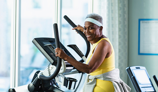 A senior African-American woman, in her 70s, at the gym exercising on a stepper or stair climbing machine. She is smiling at the camera.