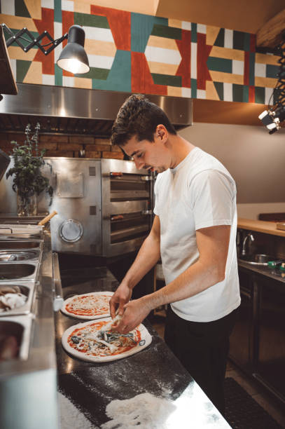 istock Young caucasian man working at pizzeria adding toppings to pizzas 1390928758