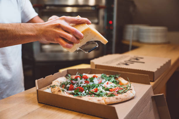 Persons hands grating parmesan cheese on freshly baked pizza in a takeaway box.