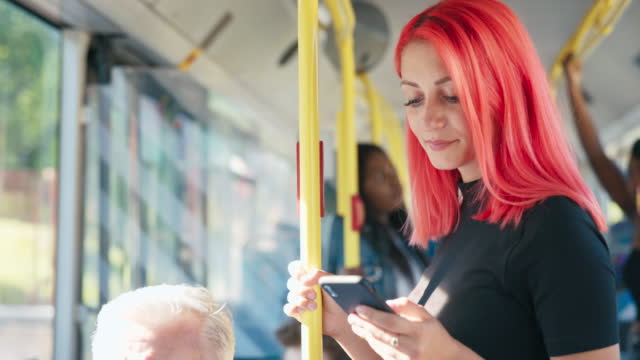 A beautiful smiling girl with pink hair rides the bus holding on to the railing