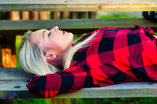 Serene young woman enjoying a sleep or nap on a park bench lying back with hands behind her head in a colorful red shirt in a close up side view