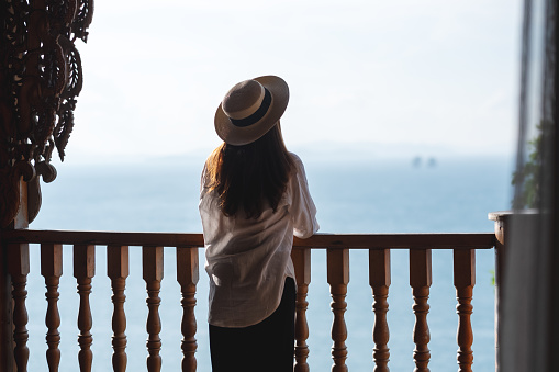 Rear view image of a young asian woman with hat looking at a beautiful sea view from resort terrace