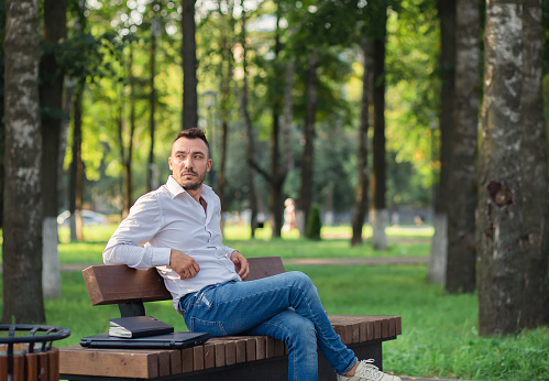 A business handsome man in a white shirt is resting sitting on a bench. Young man in city park, warm sunny summer day. Warm soft light, general plan.