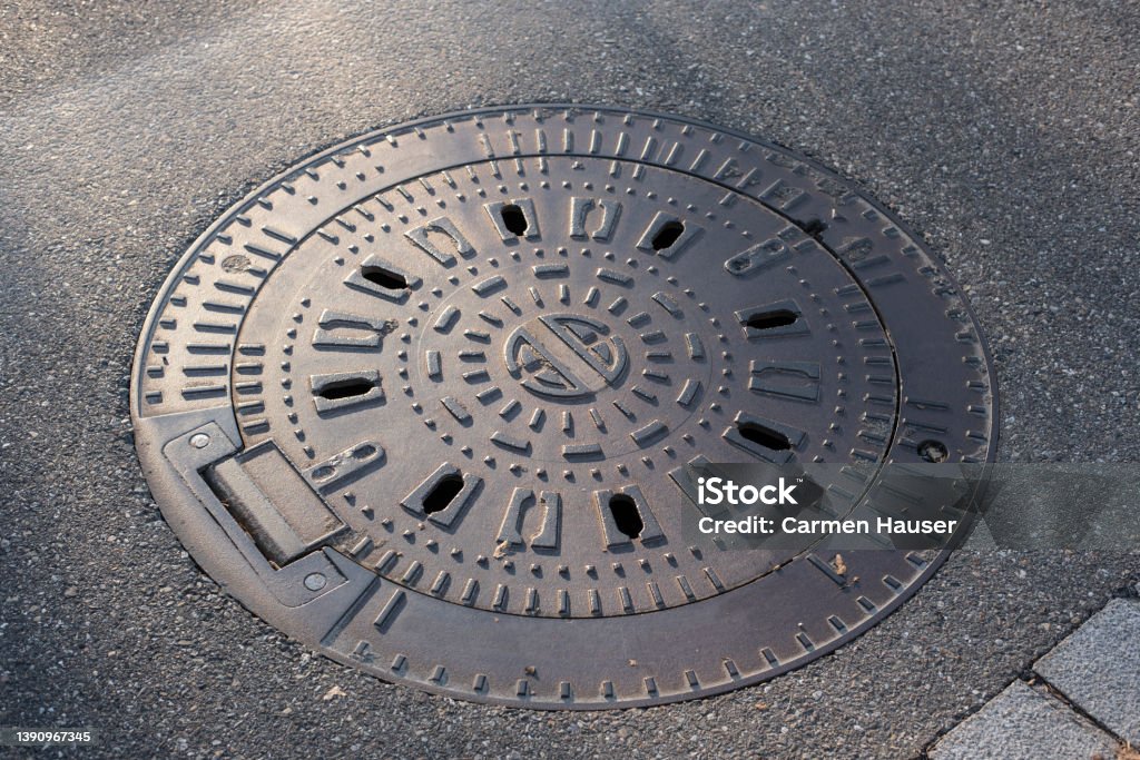 the iron cover of a manhole in a street sideview of the iron cover of a watertight manhole in a city street in evening sunlight Manhole Stock Photo
