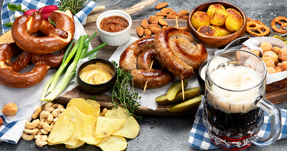 Octoberfest dinner table concept with grilled pork sausages, pretzel pastry, potatoes, salad, snacks, sauces, beer on dark background, top view.