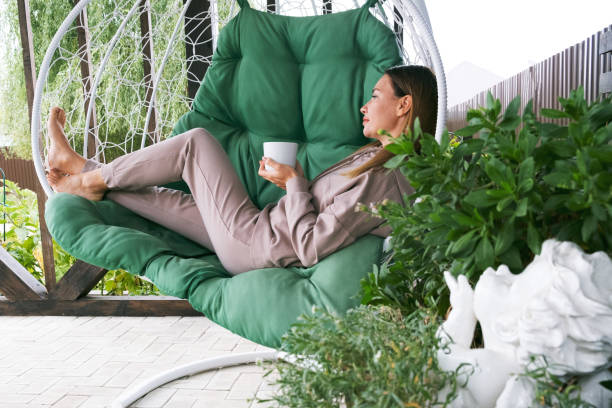Real woman drinking tea or coffee outdoor. full length mature woman sitting in a hammock chair with a cup of hot drink and relaxing in nature. close eyes. green plant. outdoor hammock. woman chilling enjoy relaxing sitting in fabric chair hammock at yard in garen near house. Peaceful and idyllic outdoor rest lifestyle mujer real bebiendo té o café al aire libre. mujer madura de cuerpo entero sentada en una silla de hamaca con una taza de bebida caliente y relajante en la naturaleza. ojos cerrados. planta verde. hamaca al aire libre. mujer escalofriante disfrutar de l