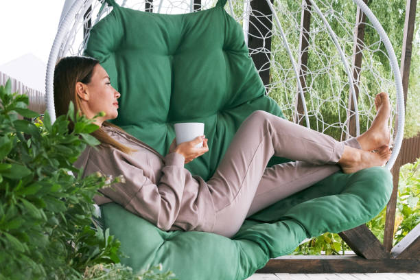 야외에서 차나 커피를 마시는 진짜 여자. 전체 길이의 성숙한 여성은 뜨거운 음료 한 잔과 함께 해먹 의자에 앉아 자연에서 휴식을 취합니다. 눈을 감으십시오. 녹색 식물. 야외 해먹.  차가워지� - summer women hammock nature 뉴스 사진 이미지