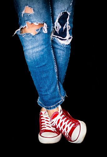 Legs Of A Person In Ripped Jeans And Red Casual Sneakers On A Black Background.  Fashion Concept. Unrecognizable Person