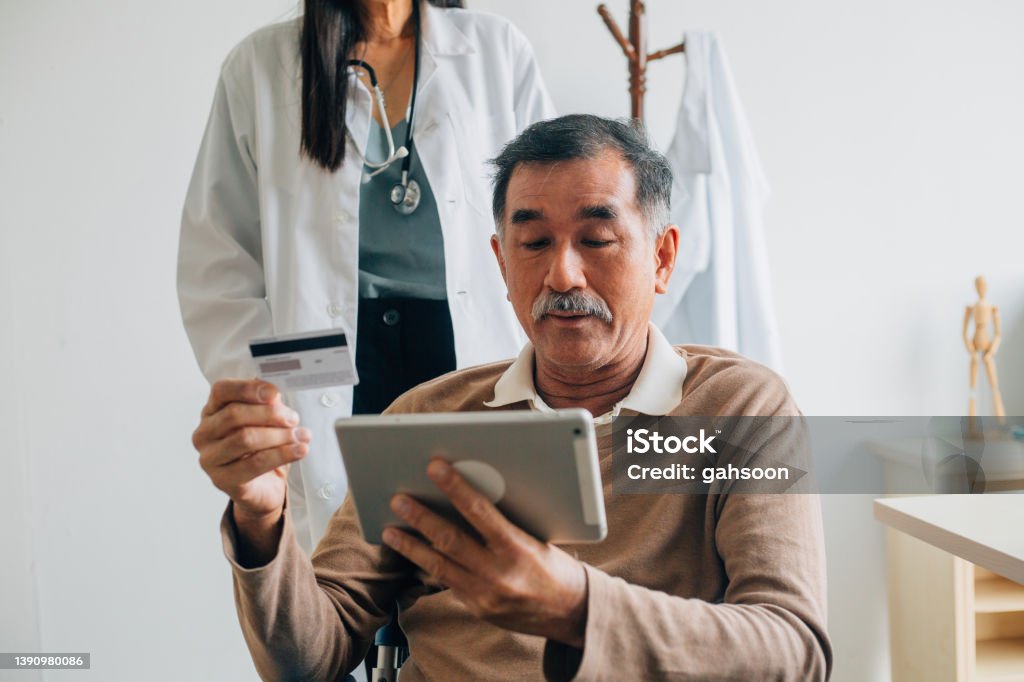 Senior patient making contactless payment using credit card in treatment clinic Senior patient using digital tablet and credit card making online payment while sitting on wheelchair at the hospital Paying Stock Photo