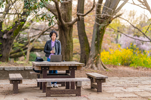A young woman is standing and using a laptop and listening to music in a public park. She is using a reusable water bottle.