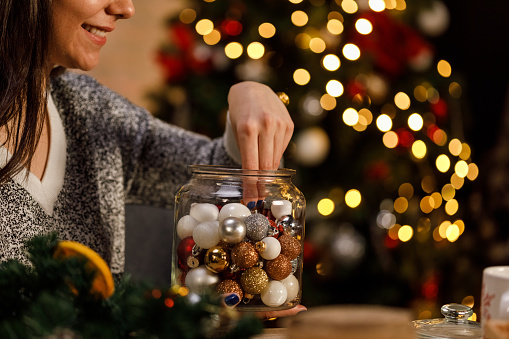 Copy space shot of smiling young woman sitting at table and picking Christmas ornaments from a glass jar, to put as a decoration on a Christmas wreath she is making.
