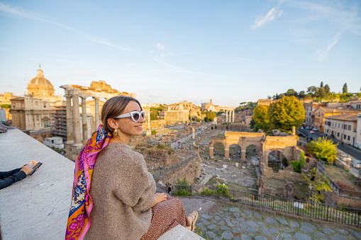 Woman enjoying view on the Roman Forum, ruins at the center of Rome on a sunset. Concept of traveling famous landmarks in Italy. Caucasian woman wearing colorful shawl in hair and sunglasses