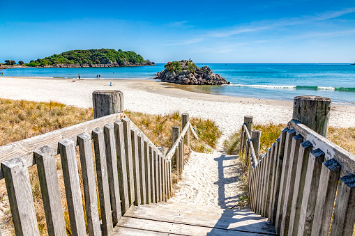 Mount Maunganui, Bay of Plenty, New Zealand - Steps down to beach on a beautiful summer day, with a small island and people walking at the edge of the sea.