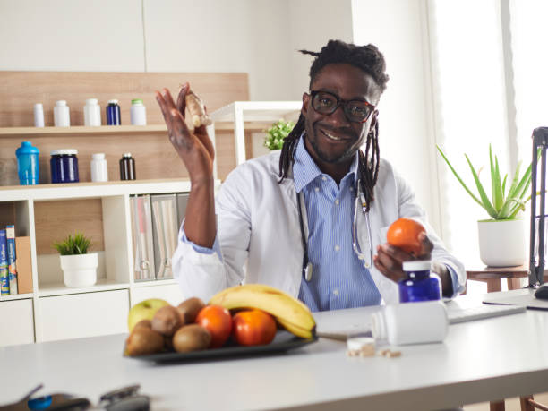 nutrizionista afroamericano che guarda la fotocamera e mostra frutti sani nella consultazione. - orange uniform foto e immagini stock