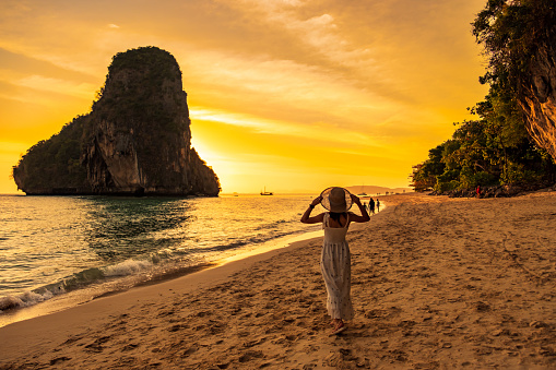 Woman tourist in white dress and hat walking on Phra nang Cave Beach at sunset, Railay, Krabi, Thailand. vacation, travel, summer, Wanderlust and holiday concept