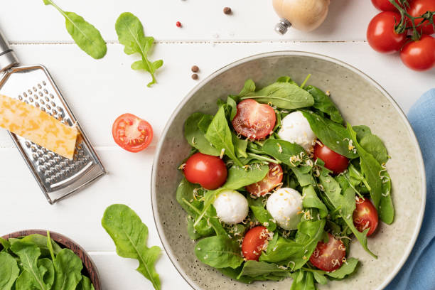 istock Fresh salad with arugula, cherry tomatoes, mozzarella cheese and hard cheese on white wooden background. Top view 1390993029