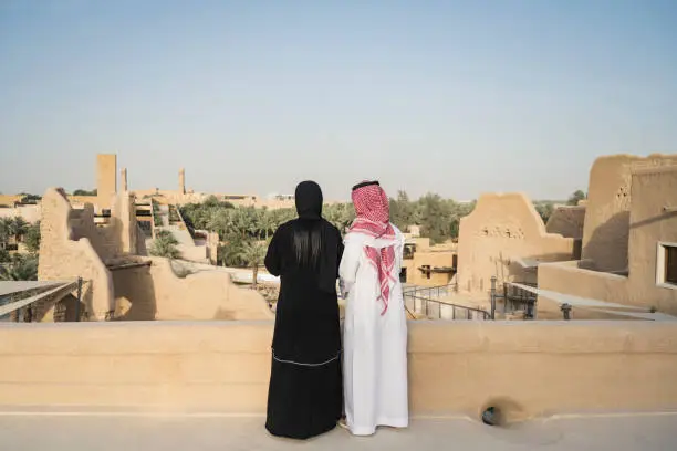 Middle Eastern woman in abaya and hijab standing with man in dish dash, kaffiyeh, and agal enjoying view of open air museum and desert oasis. Property release attached.
