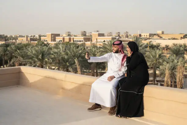 Couple in traditional Saudi attire sitting side by side on wall and smiling at smart phone as they capture memories of visit to open air museum near Riyadh. Property release attached.
